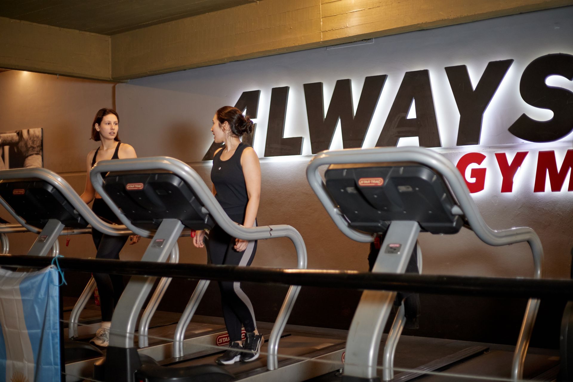 Dos mujeres en cintas de correr en un gimnasio con un letrero que dice 