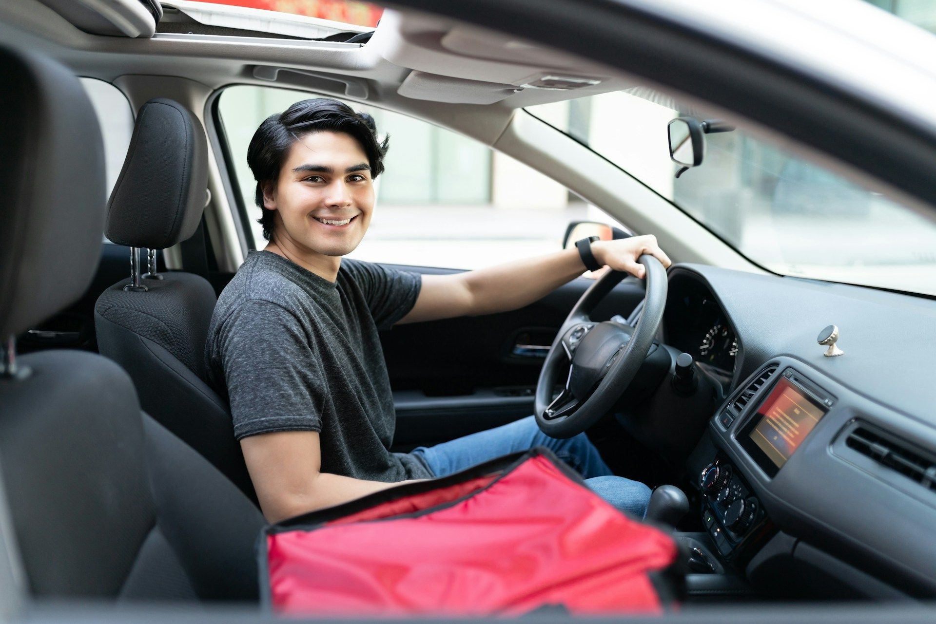Smiling driver sitting in a car with one hand on the steering wheel and a red bag in the front seat