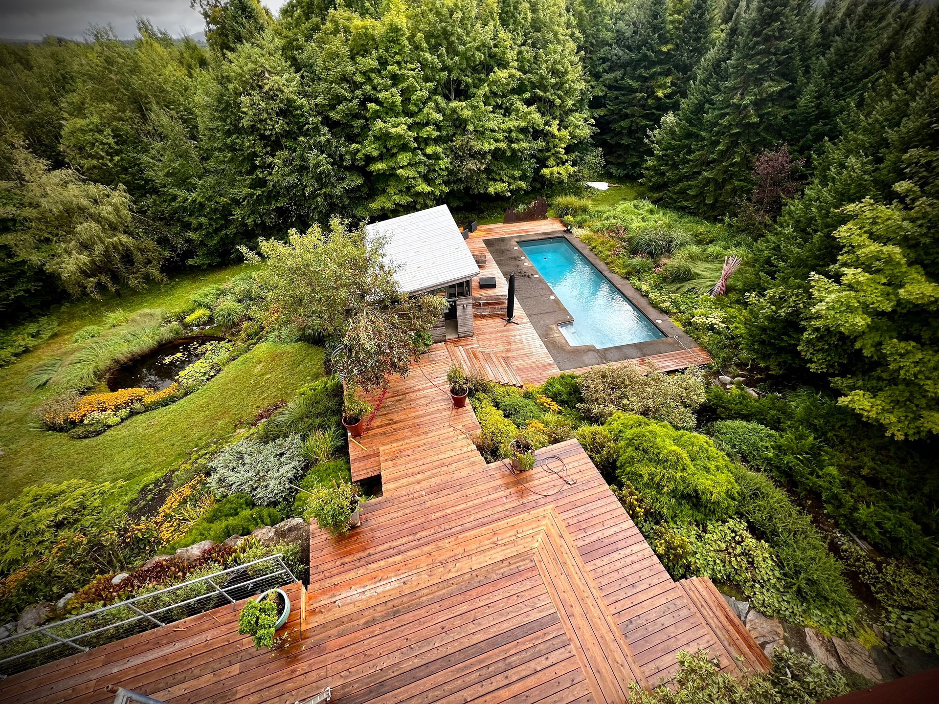 Backyard garden with wooden deck, swimming pool, and dense green trees surrounding a small patio area