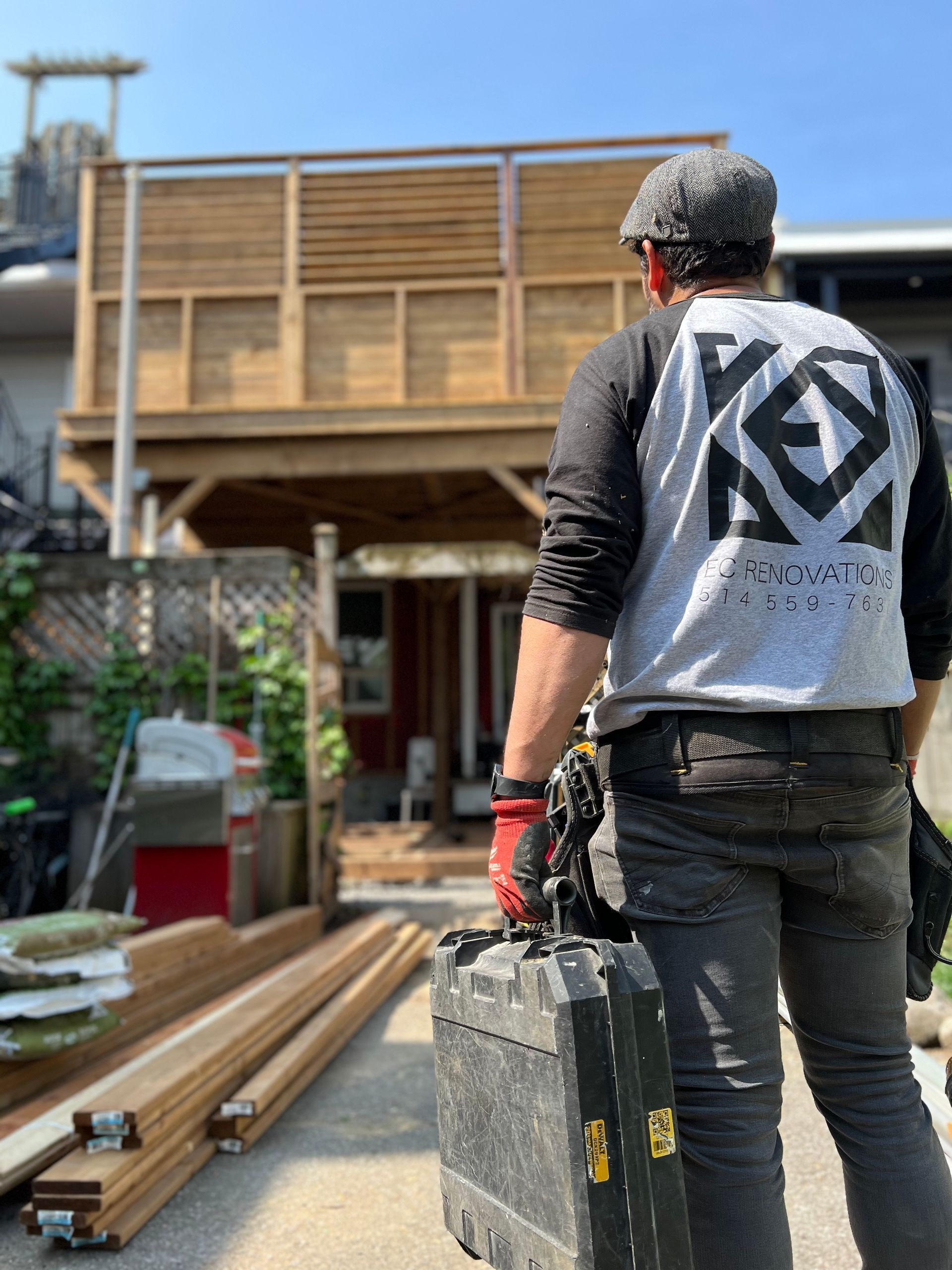 Worker in black shirt carrying a toolbox at an outdoor construction site with lumber stacked nearby