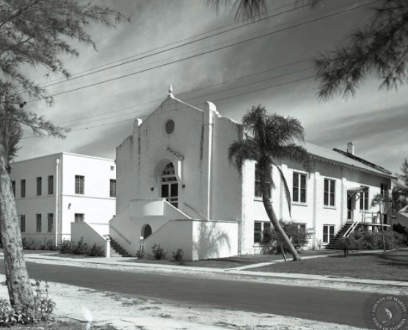 Black and white photo of a white church with a tall, central section and a lower, longer wing. Palm tree on the right.