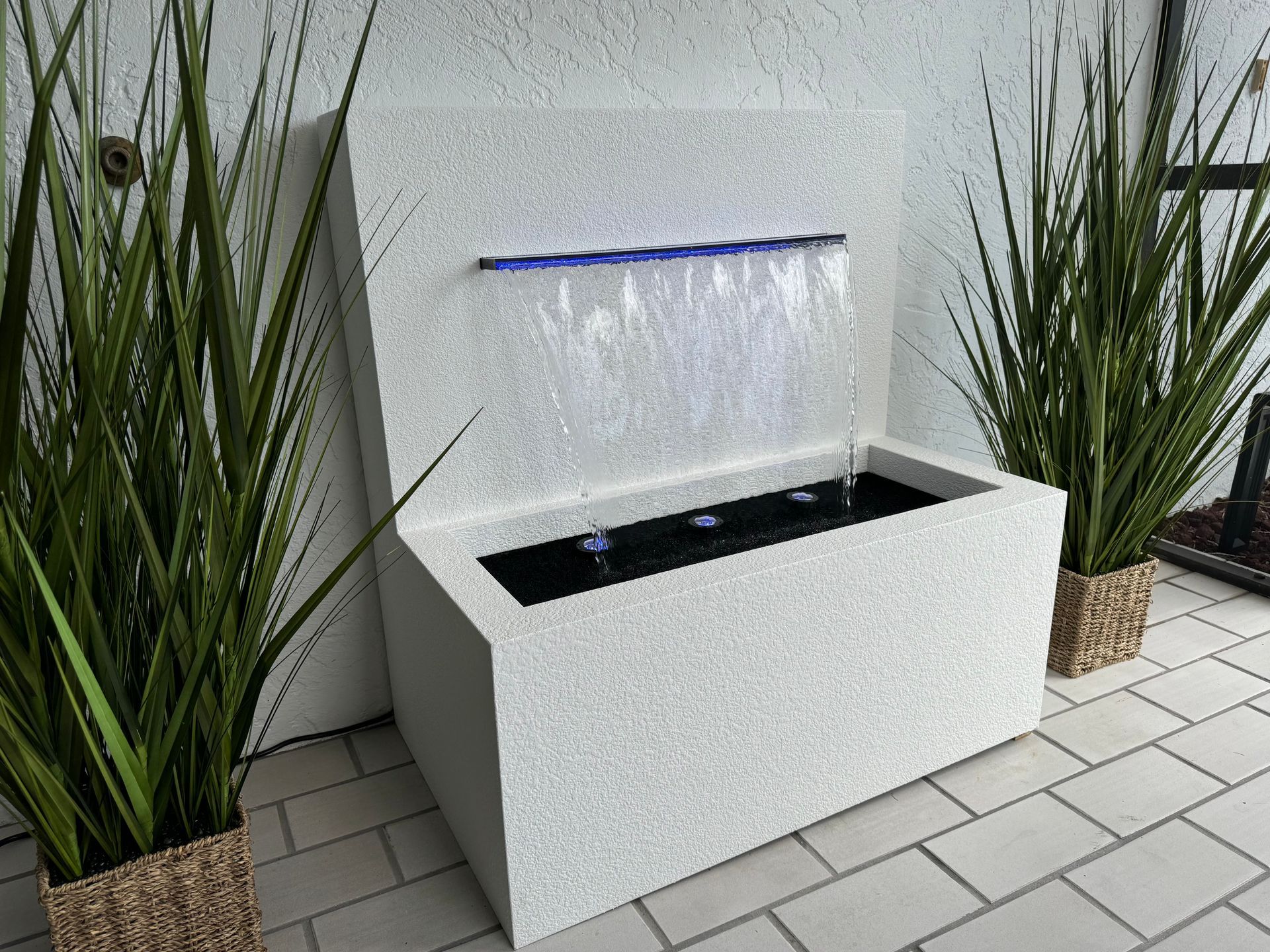 White textured water fountain with blue LED lights, flanked by potted green plants on a tiled patio