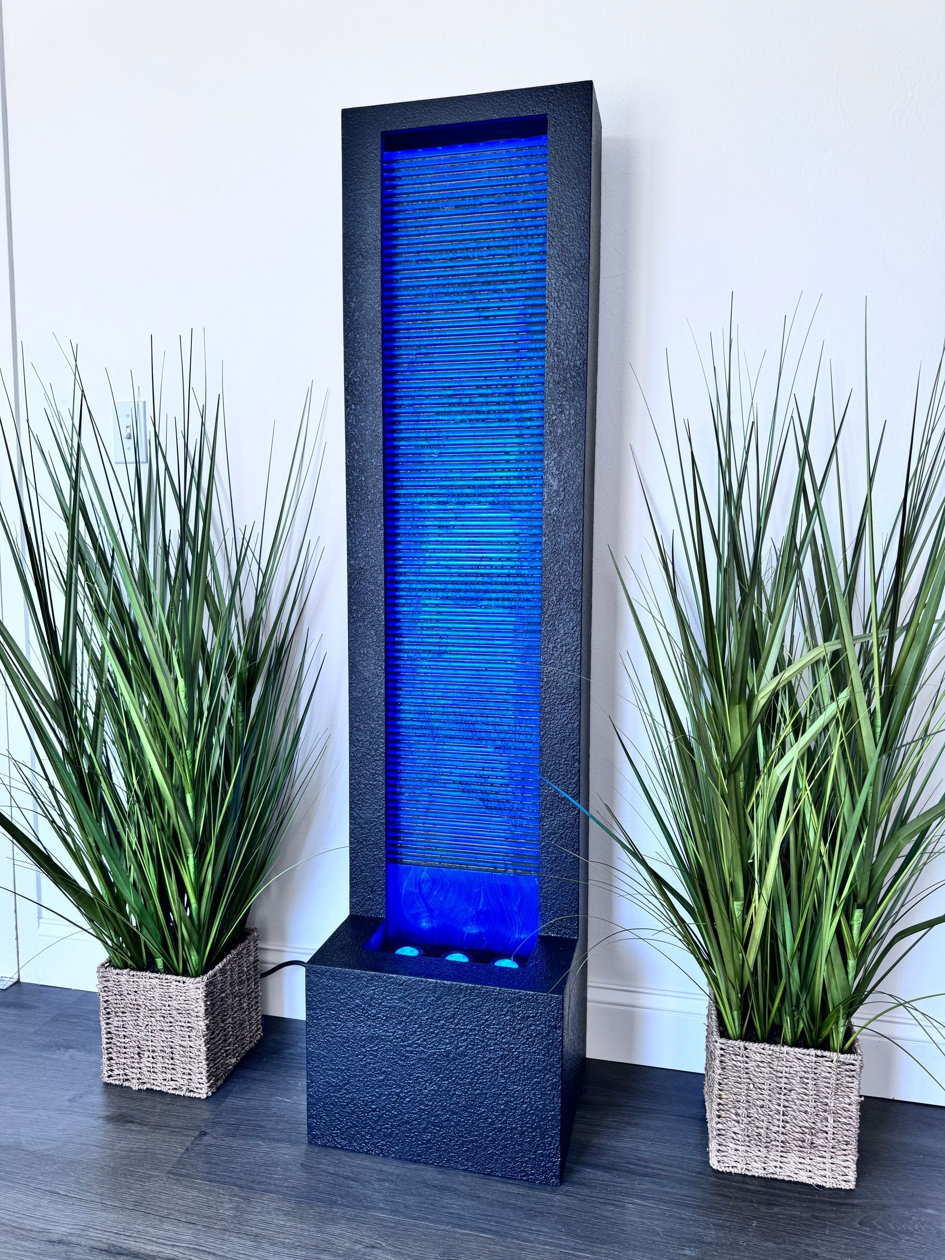 Tall, blue illuminated water fountain with black frame, flanked by potted plants