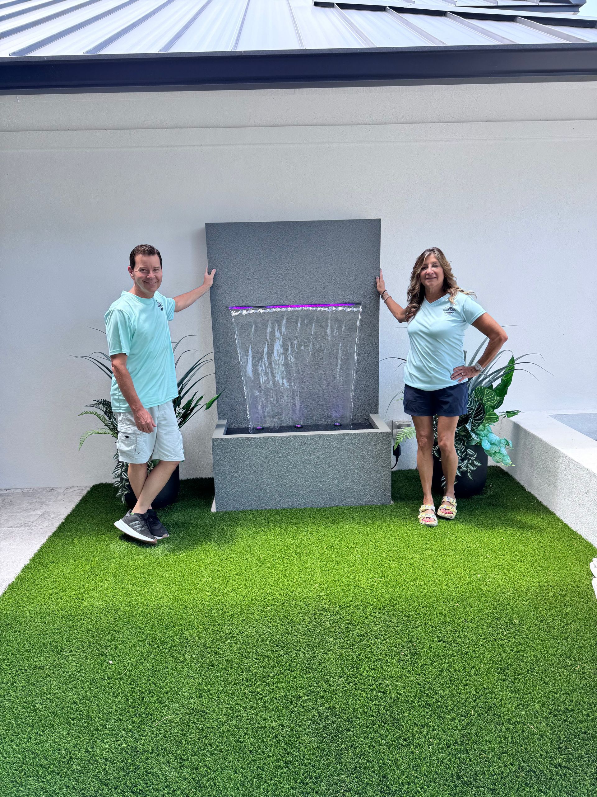 Couple poses next to a modern, gray outdoor water fountain on artificial turf