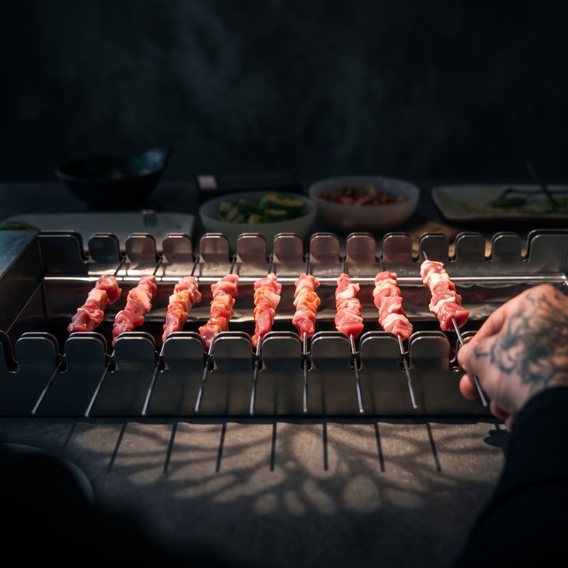 Skewers of raw meat arranged on a grill, with a hand adjusting them in dim light.
