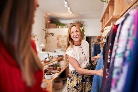 Shopkeeper smiling in a boutique, reaching toward colorful clothes on display.