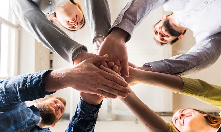 Hands stacked together in teamwork, viewed from below in a bright office setting.