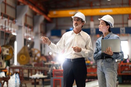 Two workers in hard hats reviewing plans in a factory aisle, one pointing ahead.