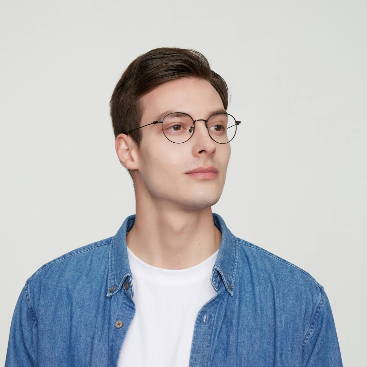 Young man in glasses wearing a blue denim shirt over a white T-shirt, looking to the side against a plain background