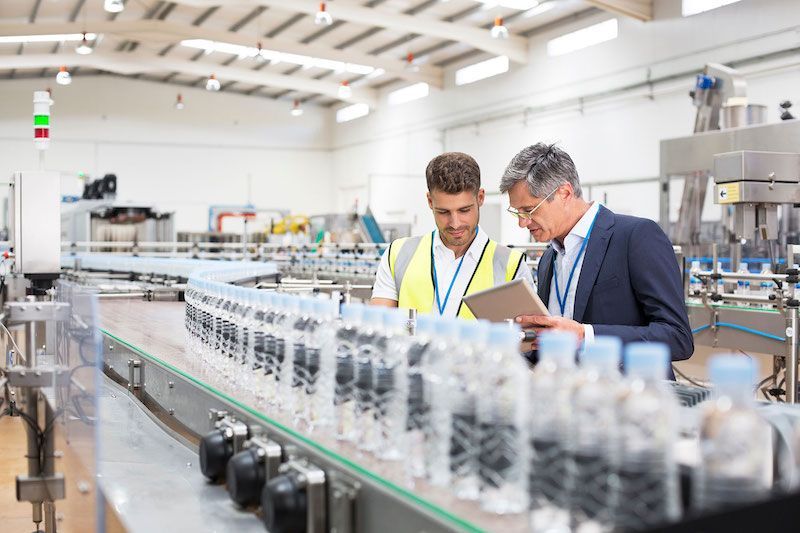 Two professionals reviewing paperwork beside a conveyor line in a bright factory.