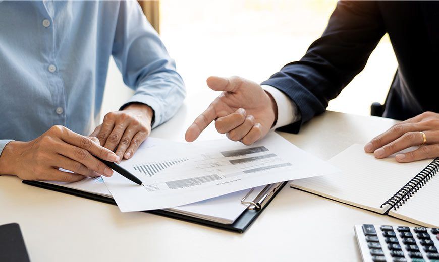 Two people reviewing documents at a desk, one pointing while the other writes notes on a clipboard.