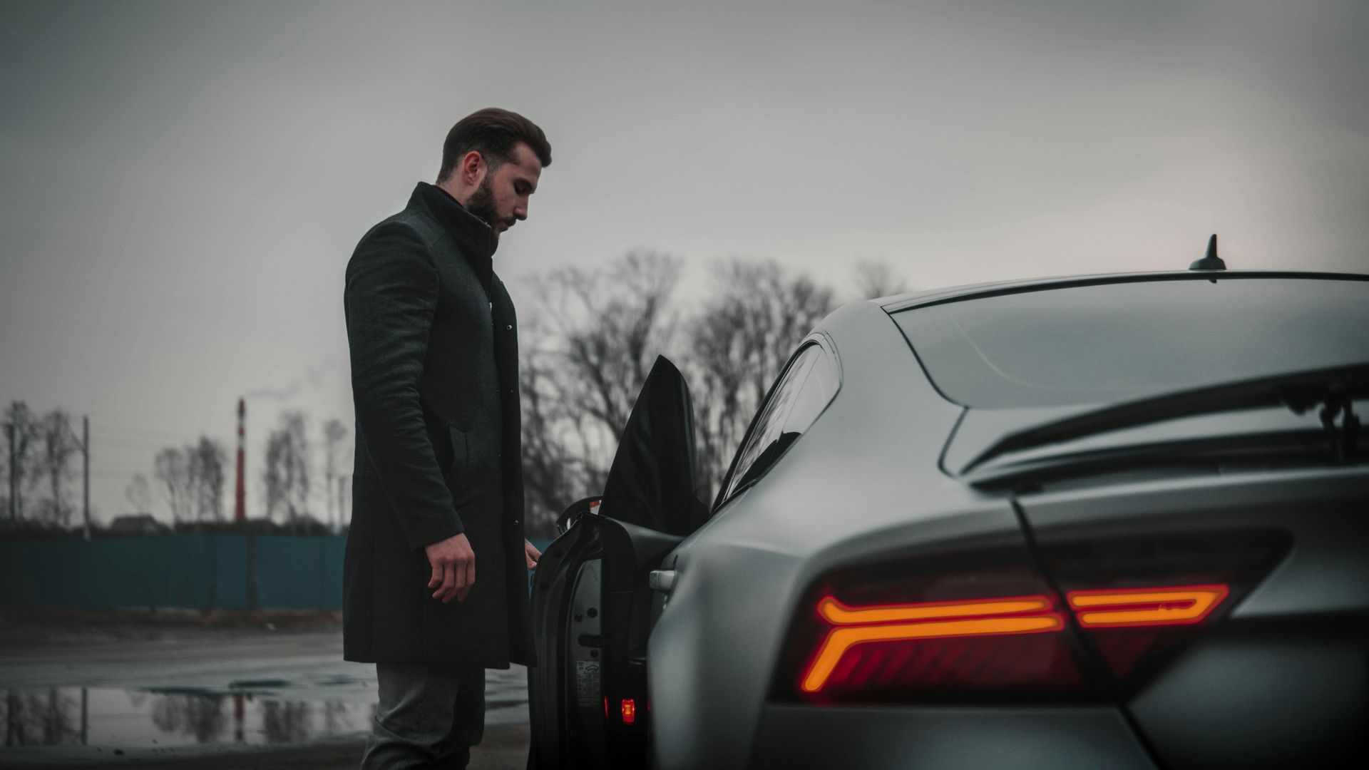 Man in black coat standing near a gray car with an open door on a cloudy day.