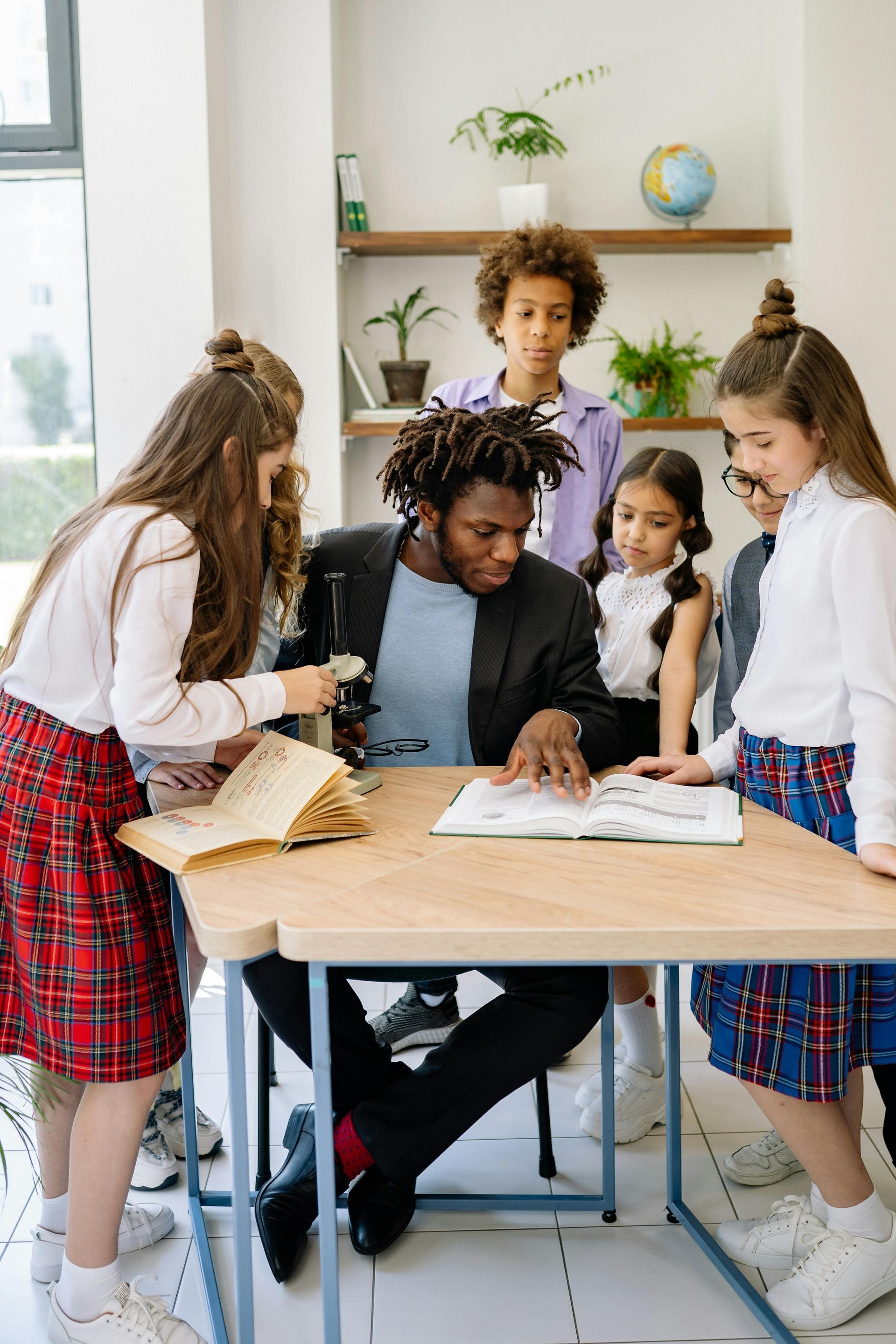 Teacher and four students gather around a table, looking at an open book in a bright classroom.
