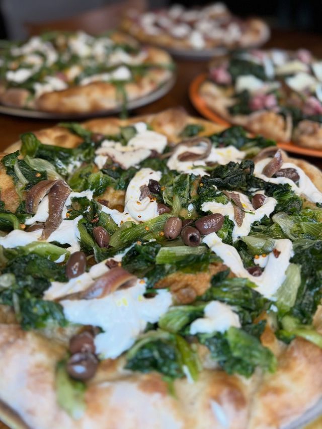 Una pizza appena sfornata condita con verdure, funghi e formaggio bianco su un tavolo, con altre pizze sullo sfondo.