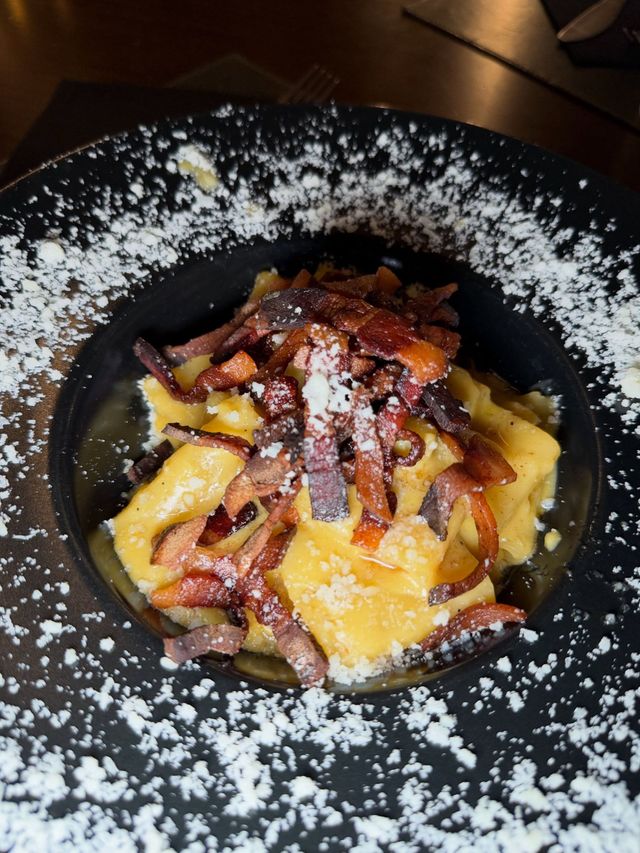 Pasta condita con pancetta su un piatto nero, cosparsa di formaggio grattugiato o sale.