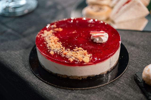 Cheesecake glassata di rosso su un piatto nero con guarnizione di briciole e una cucchiaiata di panna.