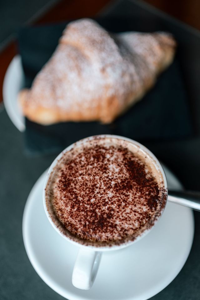 Una tazza bianca di cappuccino su un piattino, con un croissant sullo sfondo, su un tavolo scuro.