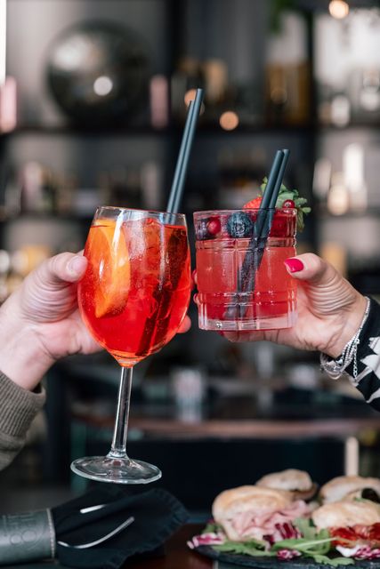 Due mani che brindano con cocktail rossi guarniti con frutti di bosco e fette d'arancia a un tavolo di un ristorante.