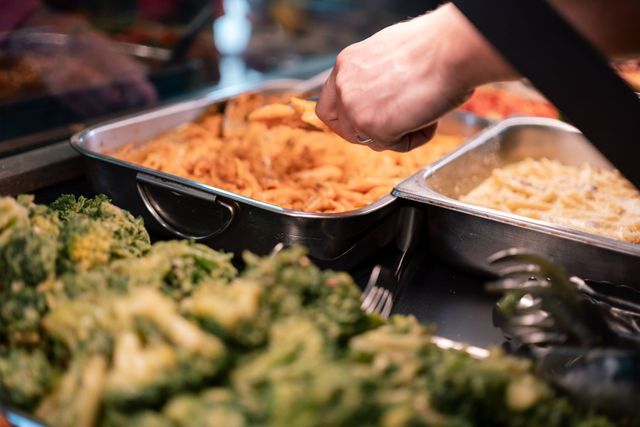 Cibo arancione cotto servito a mano da un vassoio da buffet accanto a vassoi di broccoli e pasta.