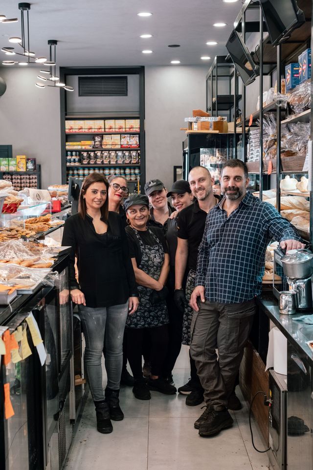 Cinque persone sono in piedi all'interno di un panificio, sorridenti accanto agli scaffali pieni di pane e pasticcini.
