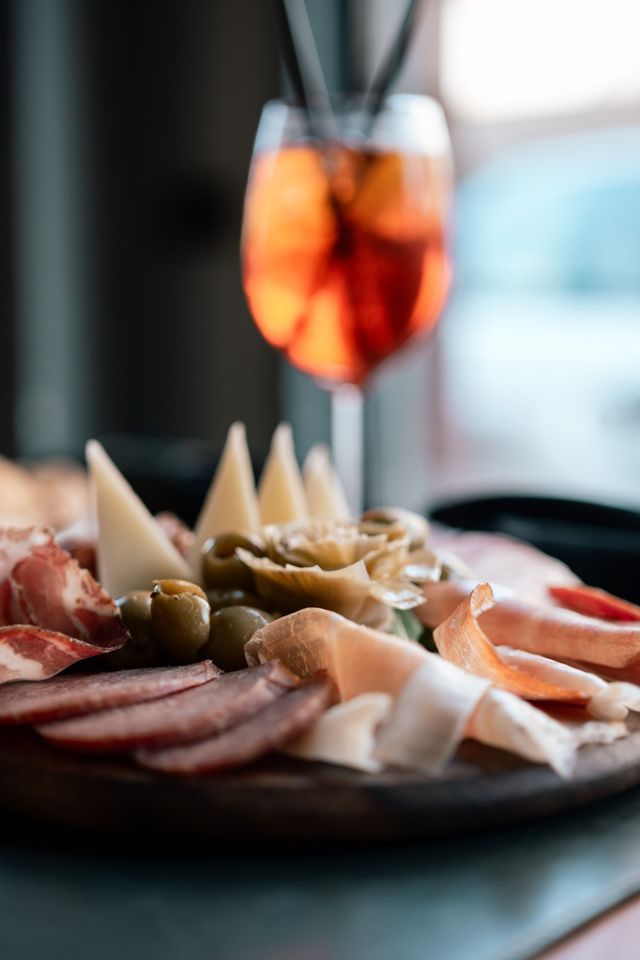 Piatto di salumi con affettati, formaggi, olive e pane, con un bicchiere di succo d'arancia sullo sfondo.