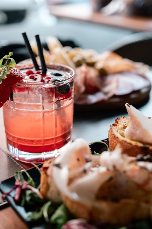 Cocktail rosa con cannucce nere accanto ad antipasti impiattati su un tavolo di ristorante.
