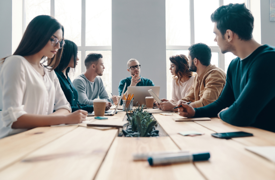 People in a bright meeting room discuss around a conference table with notebooks and coffee cups.