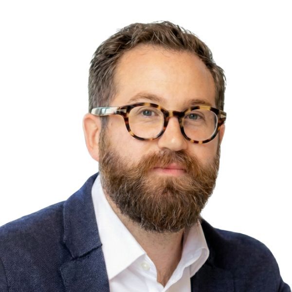 Studio headshot of a bearded man wearing glasses, a white shirt, and a navy blazer on a white background