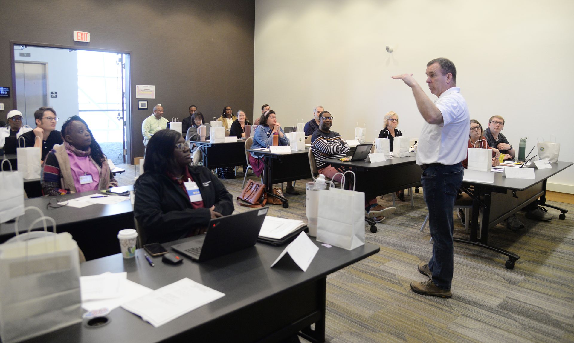 Man lecturing to a diverse group seated at desks in a conference room.