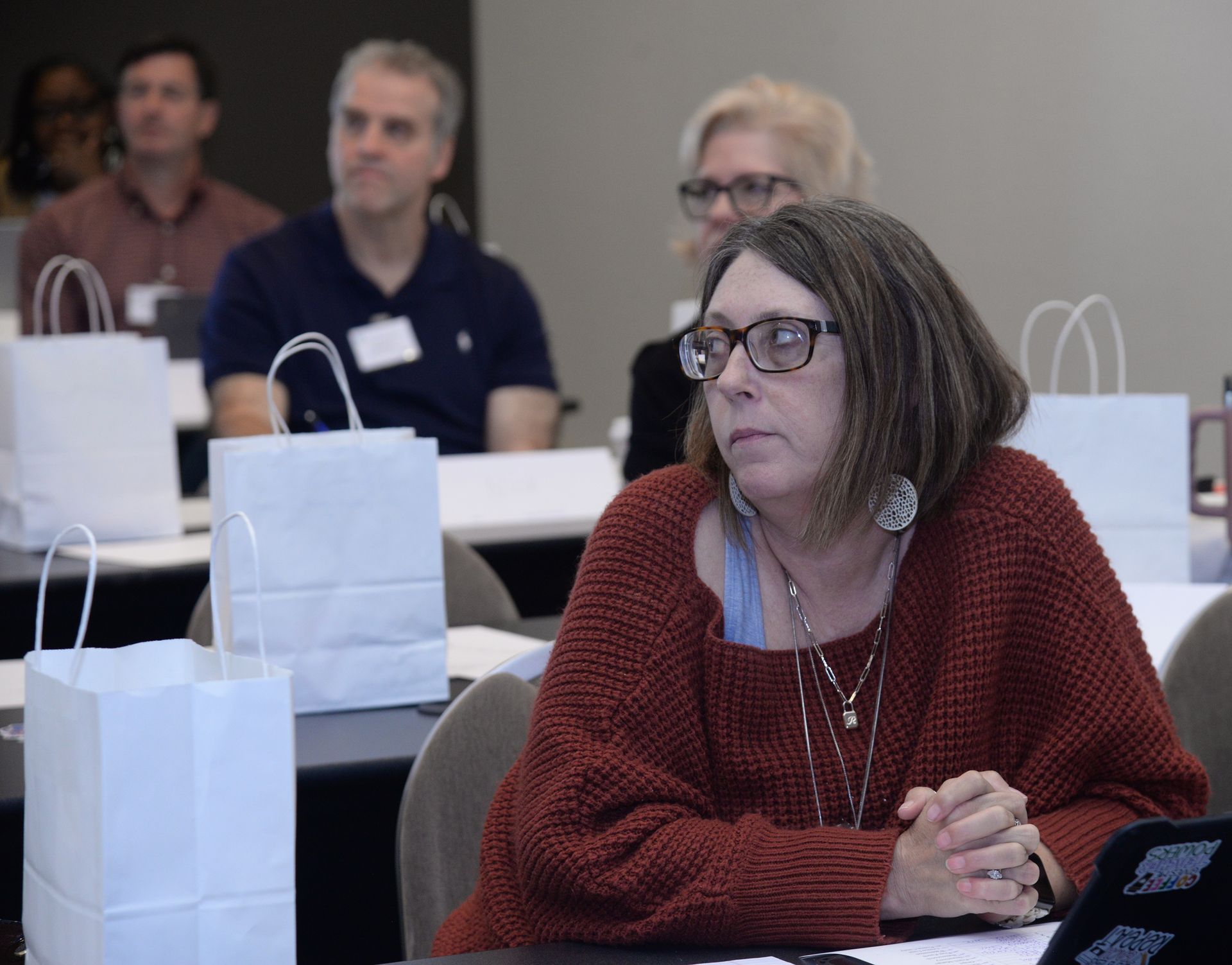 Woman in a red sweater wearing glasses and looking focused in a classroom setting, with other attendees.