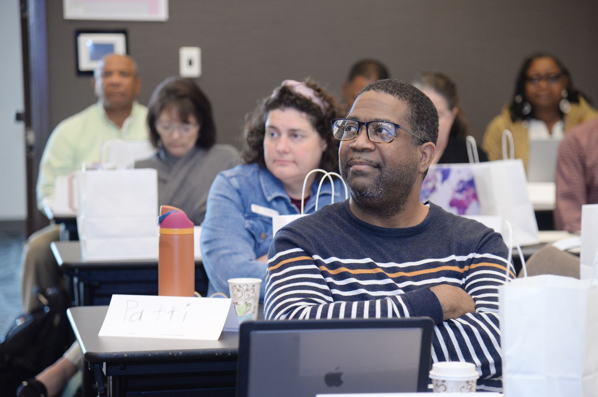 Attendees in a classroom. A man in glasses looks to the right, arms crossed. Others listen, looking forward.