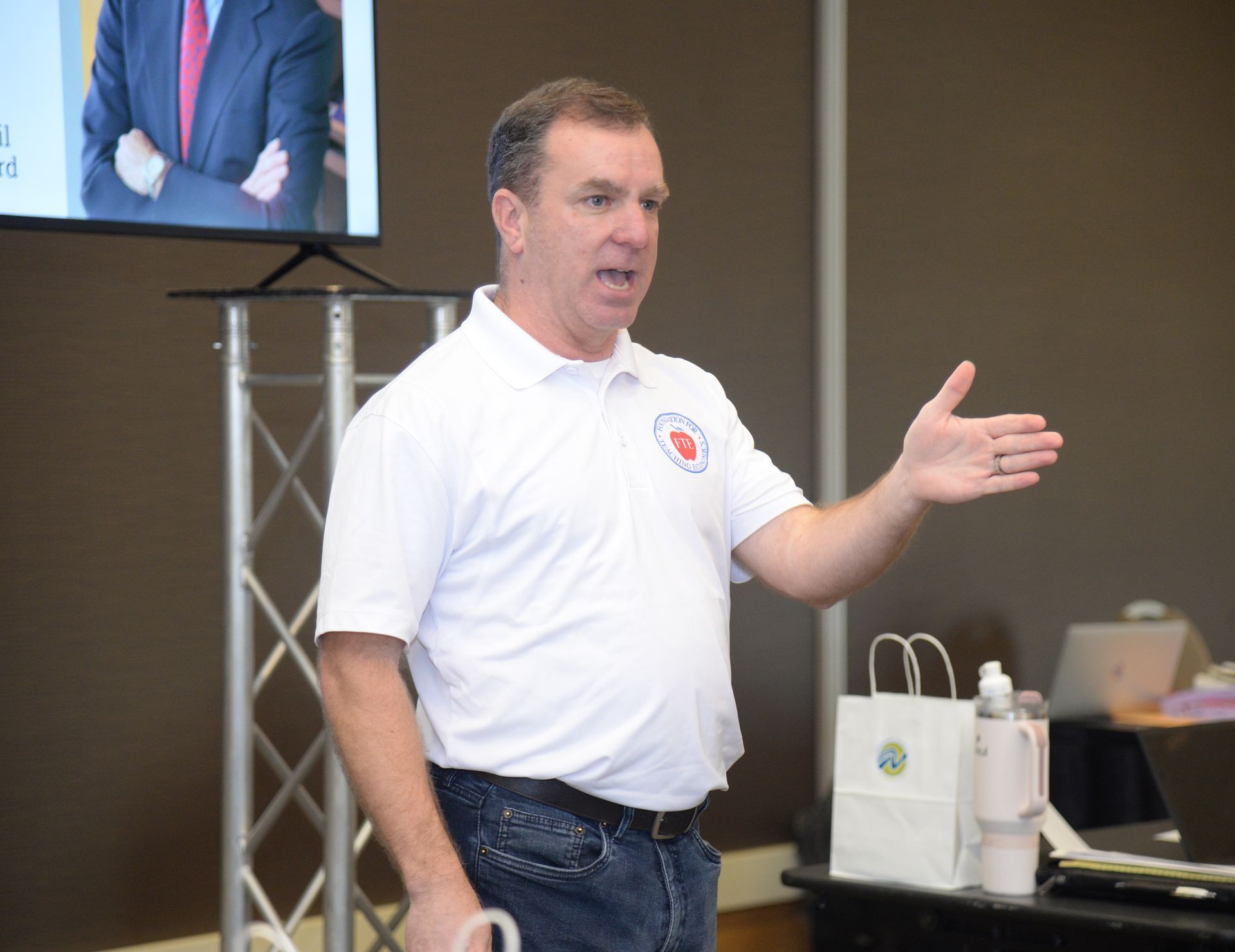 Man in white shirt gestures, speaking at a presentation; a screen, table, and bags are present.