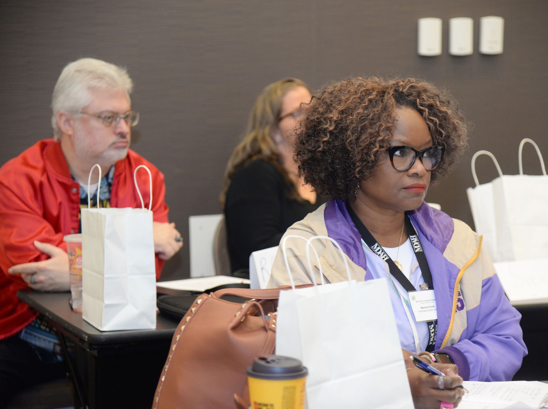 Three people seated at a table, one in red jacket, one in black, and one in a purple jacket. All looking forward.