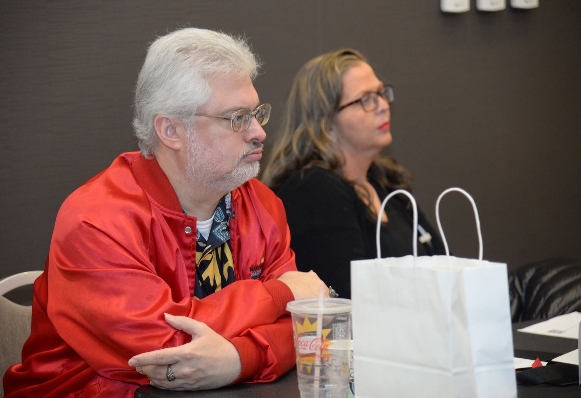 Man in red jacket and glasses, and a woman next to him looking away, both seated.
