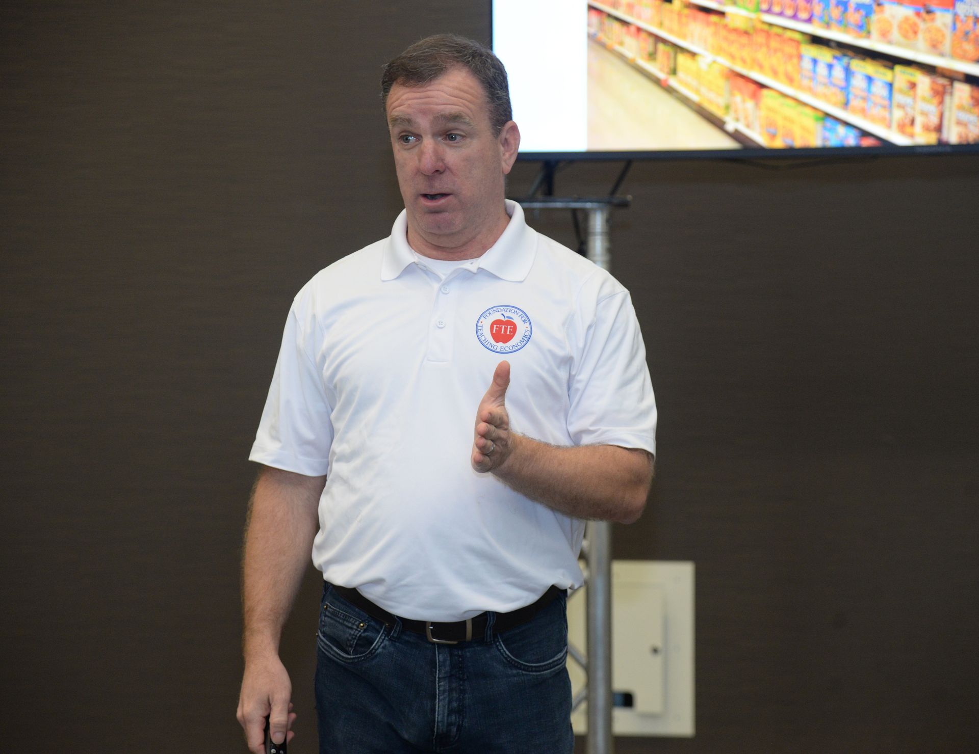 Man in white polo shirt gestures towards a screen with store shelves in the background.