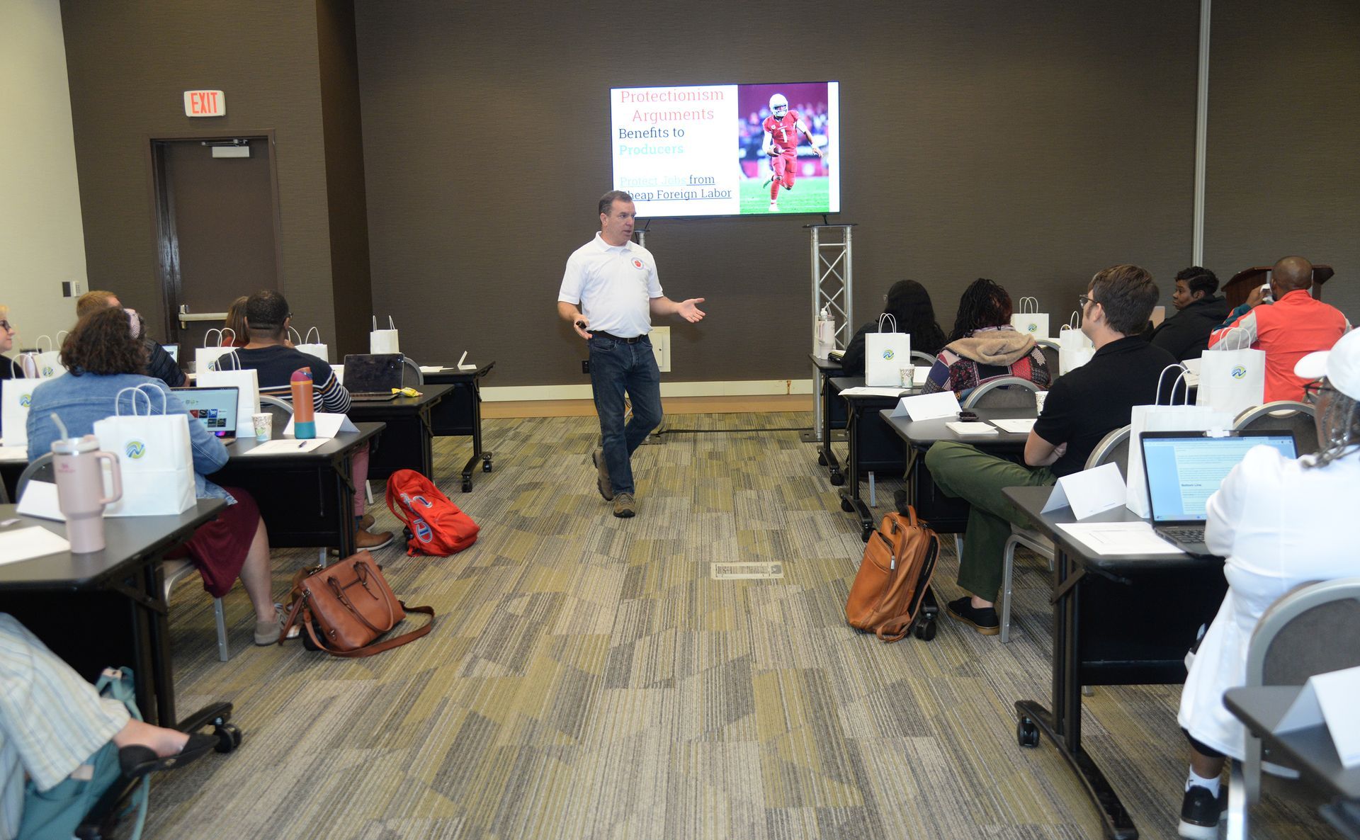 Man presenting to a group in a conference room with a screen displaying text. People are seated at desks.