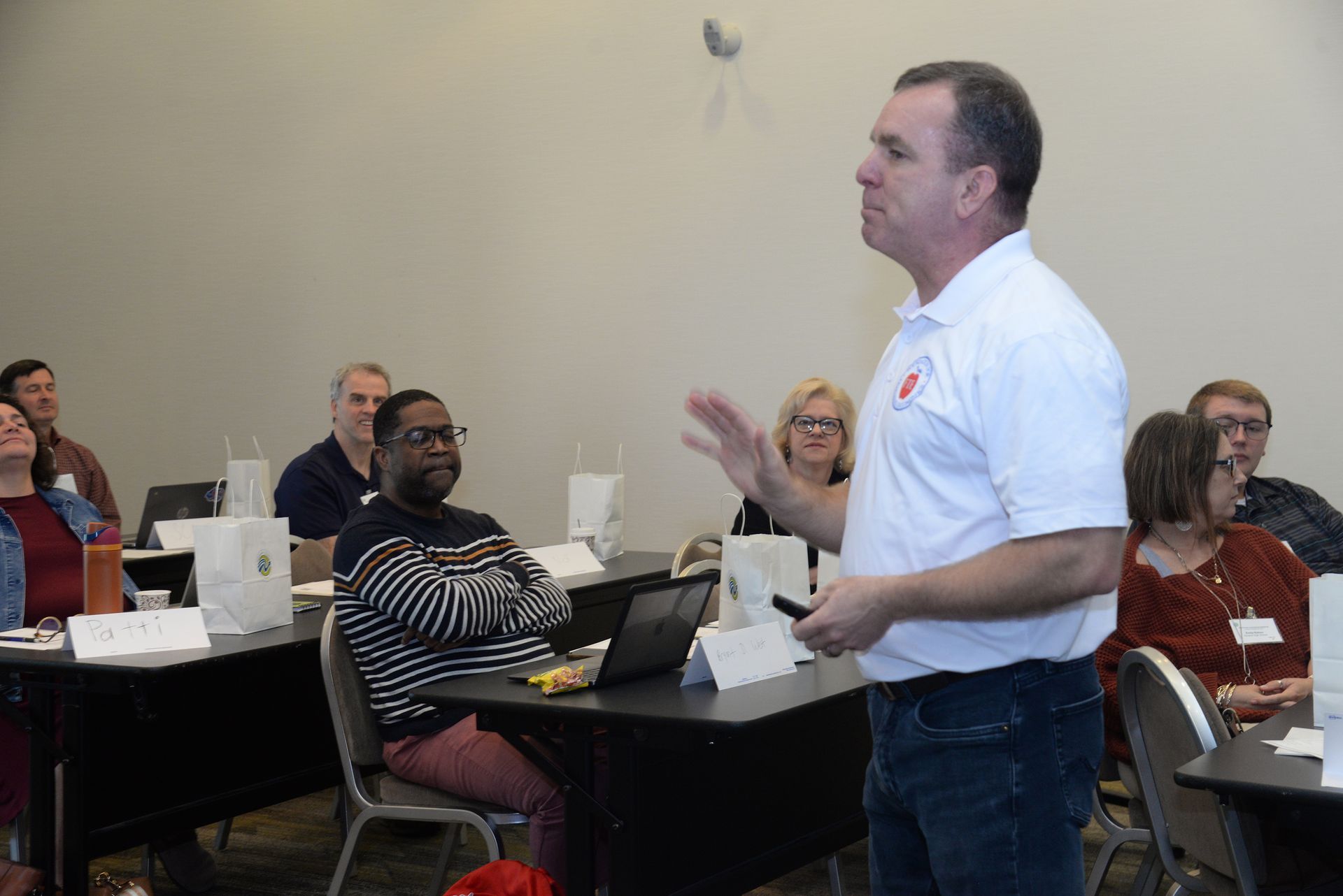 Man giving a presentation, gesturing towards the audience seated at tables.