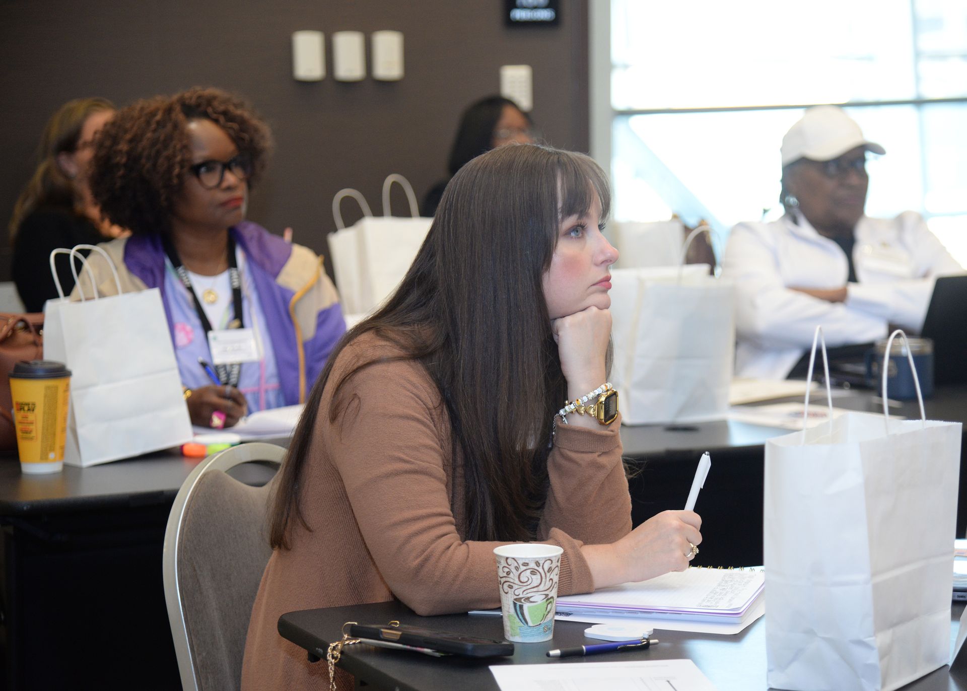 Attendees listen attentively at a workshop. A woman in a brown shirt rests her chin on her hand, holding a pen.