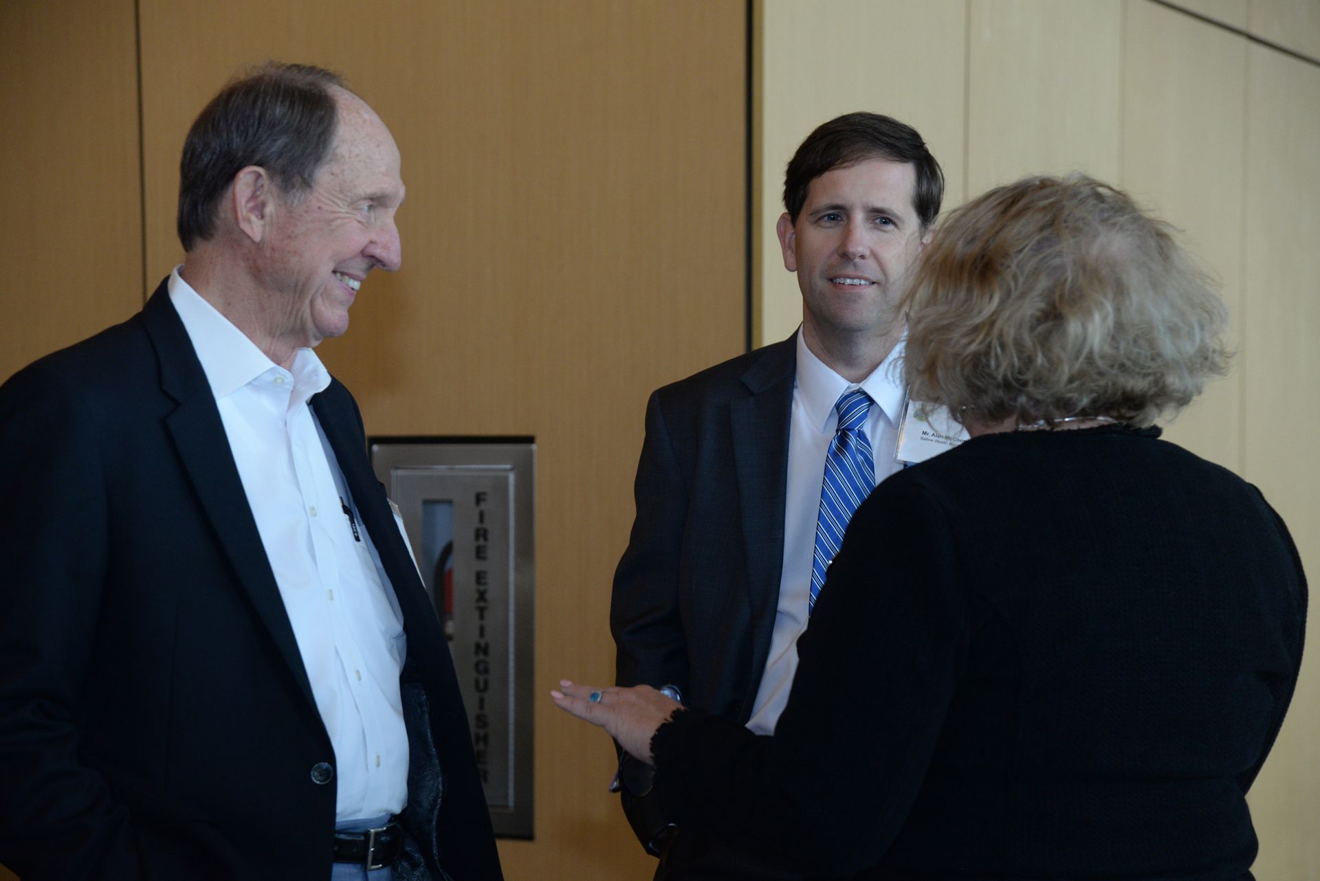 Three people in business attire conversing in an office setting; man in jacket smiles.