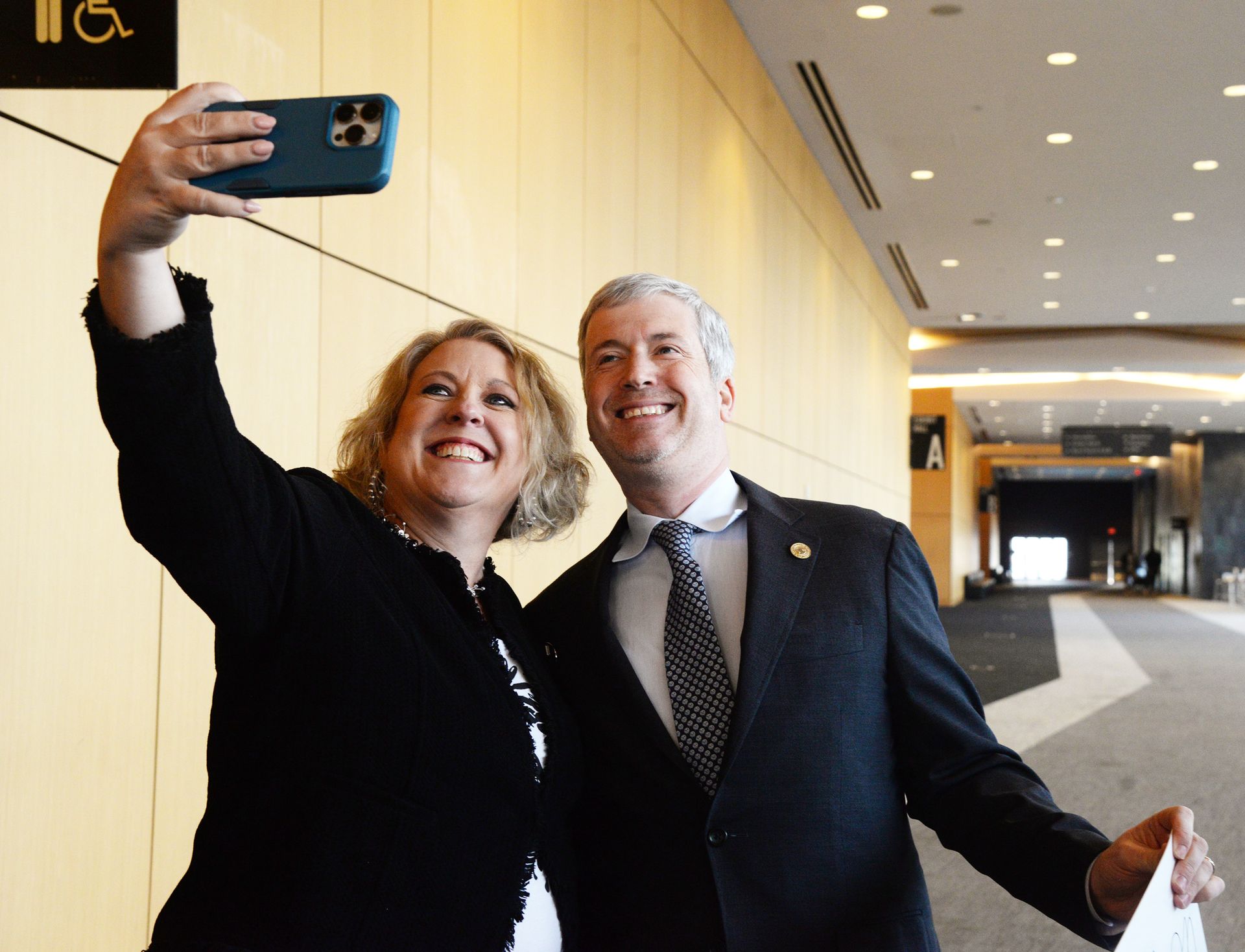 Woman taking selfie with a man in a suit. They are smiling in a modern building with beige walls.
