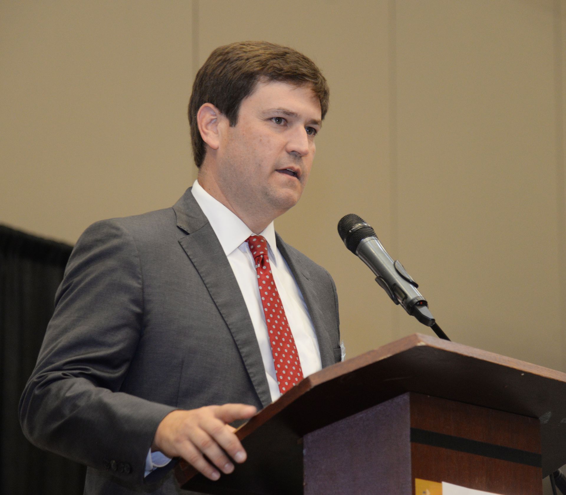 Man in gray suit speaks at a podium with a microphone. He wears a red patterned tie.