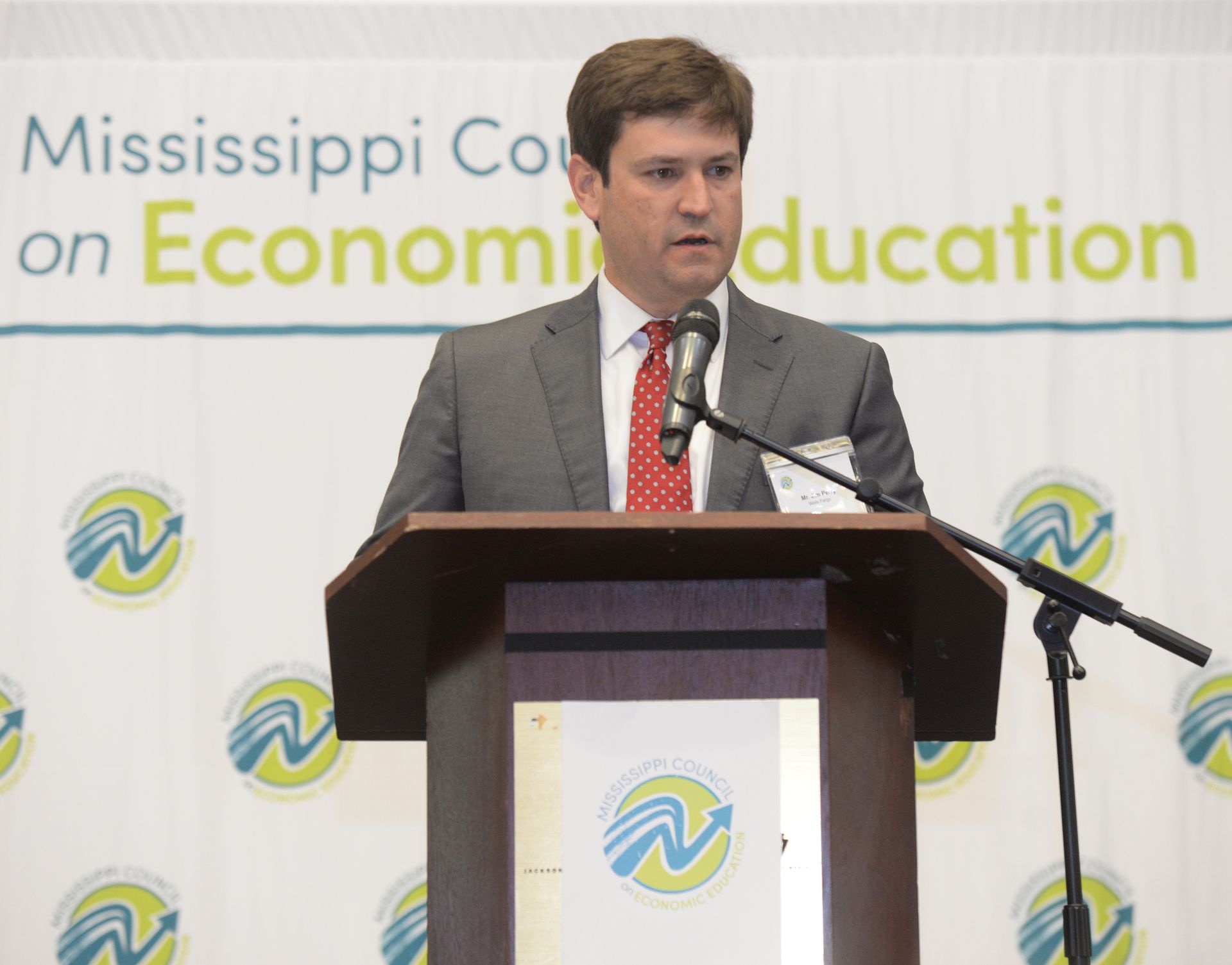 Man in a suit speaks at a podium with a microphone; background reads 
