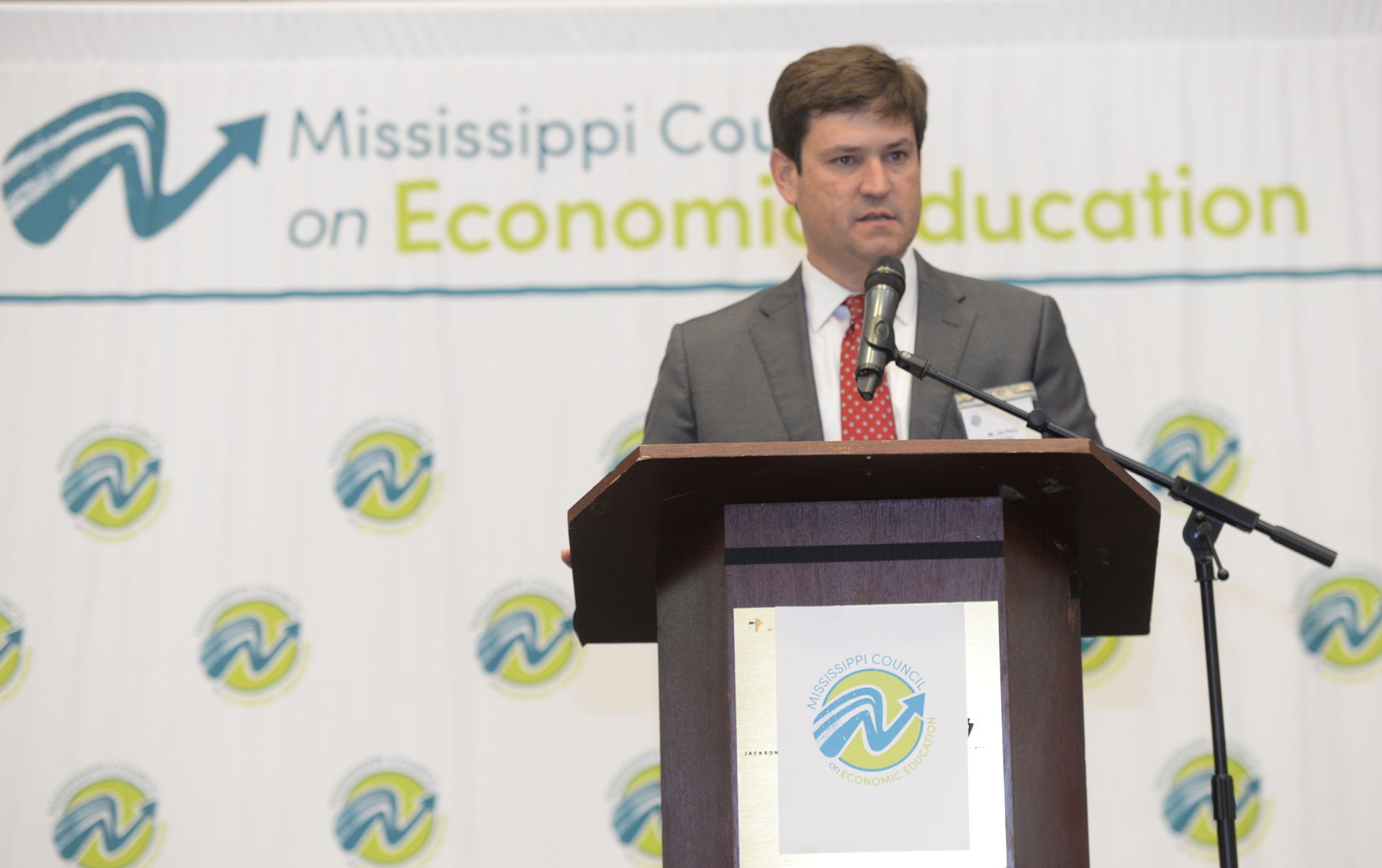 Man speaking at a podium with a microphone, Mississippi Council on Economic Education backdrop.