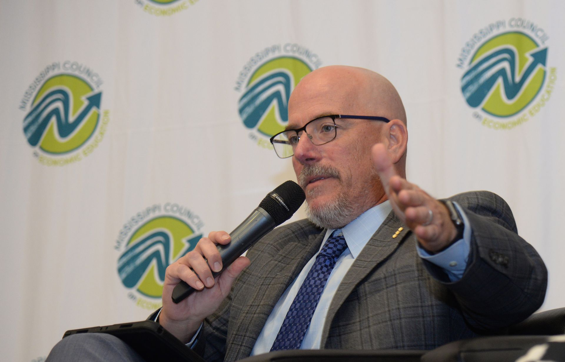 Man with glasses and beard speaks into a microphone, gesturing with his hand. Background features logos.