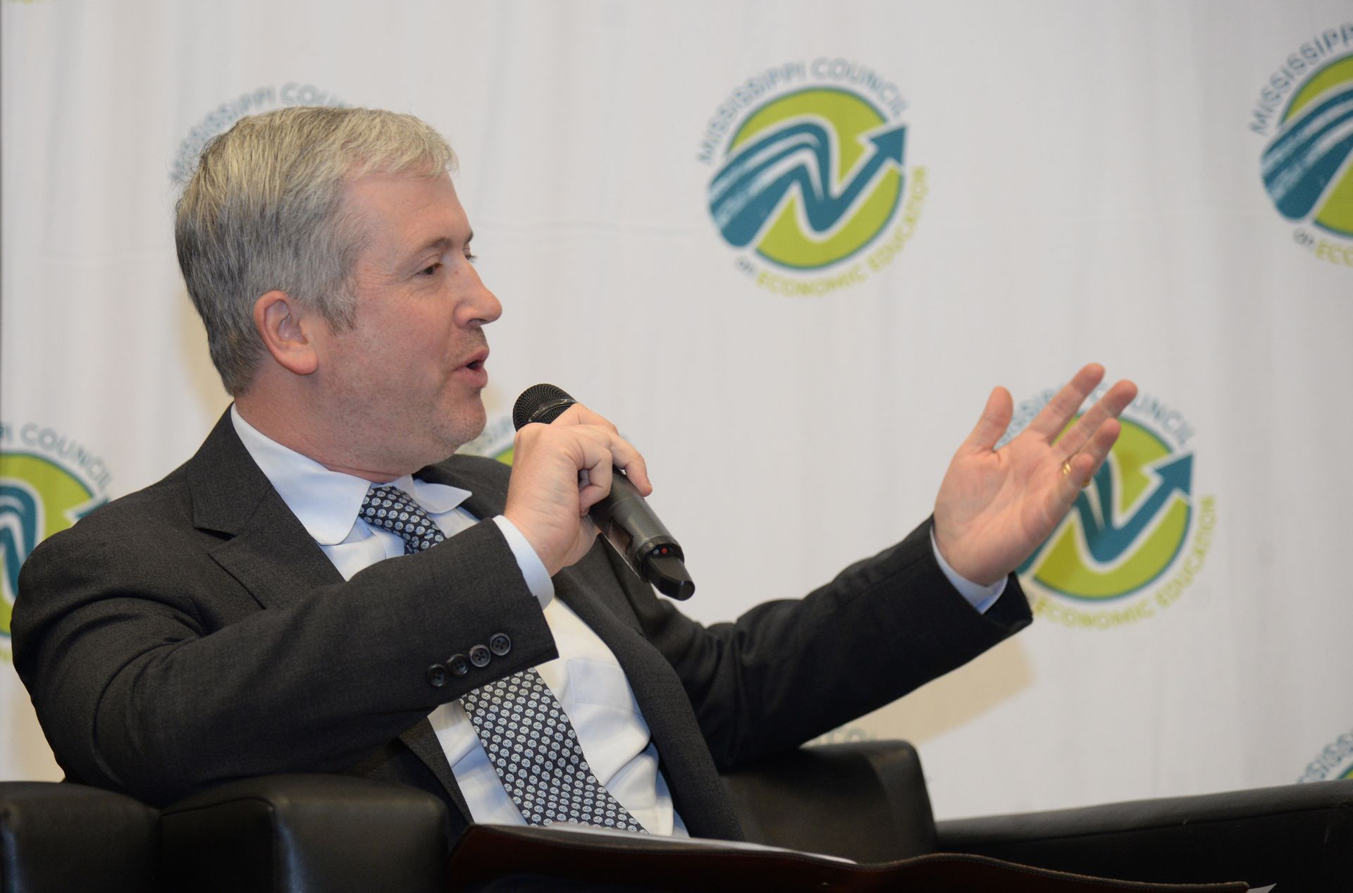Man in suit speaking into a microphone with his hand raised. Backdrop with logos.