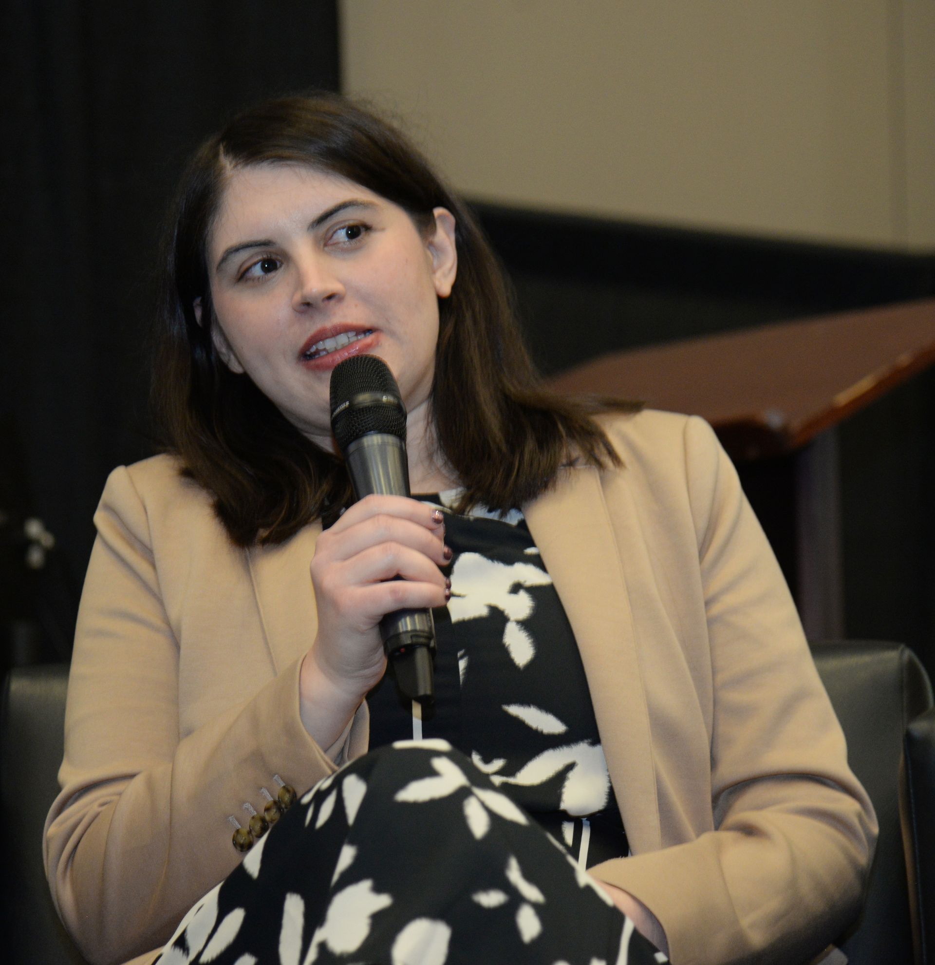 Woman speaking into a microphone, wearing a blazer and floral dress.