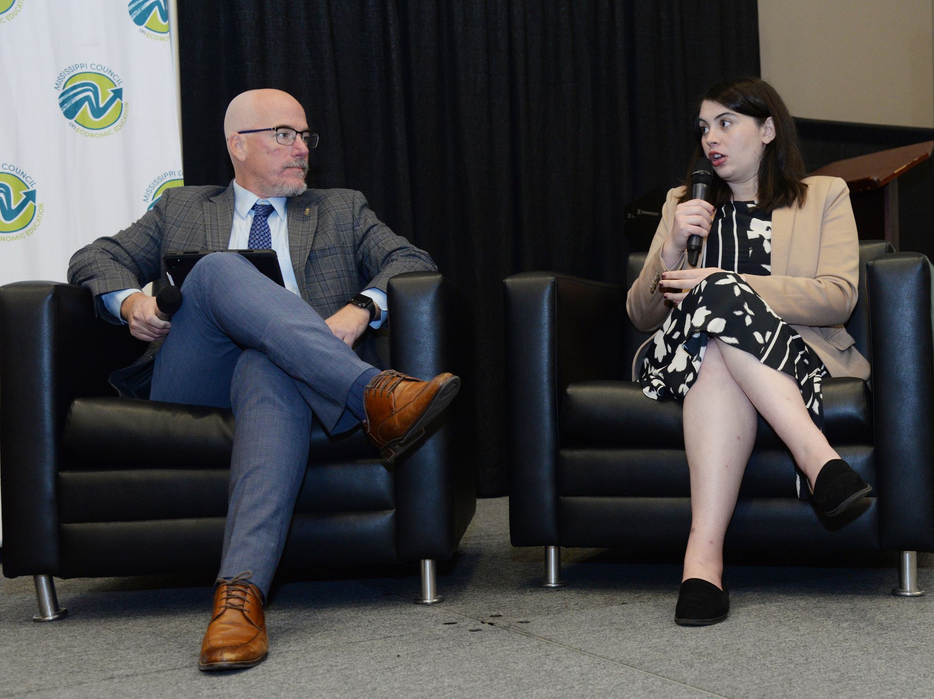 Man and woman seated in armchairs on a stage, discussing. The woman holds a microphone.