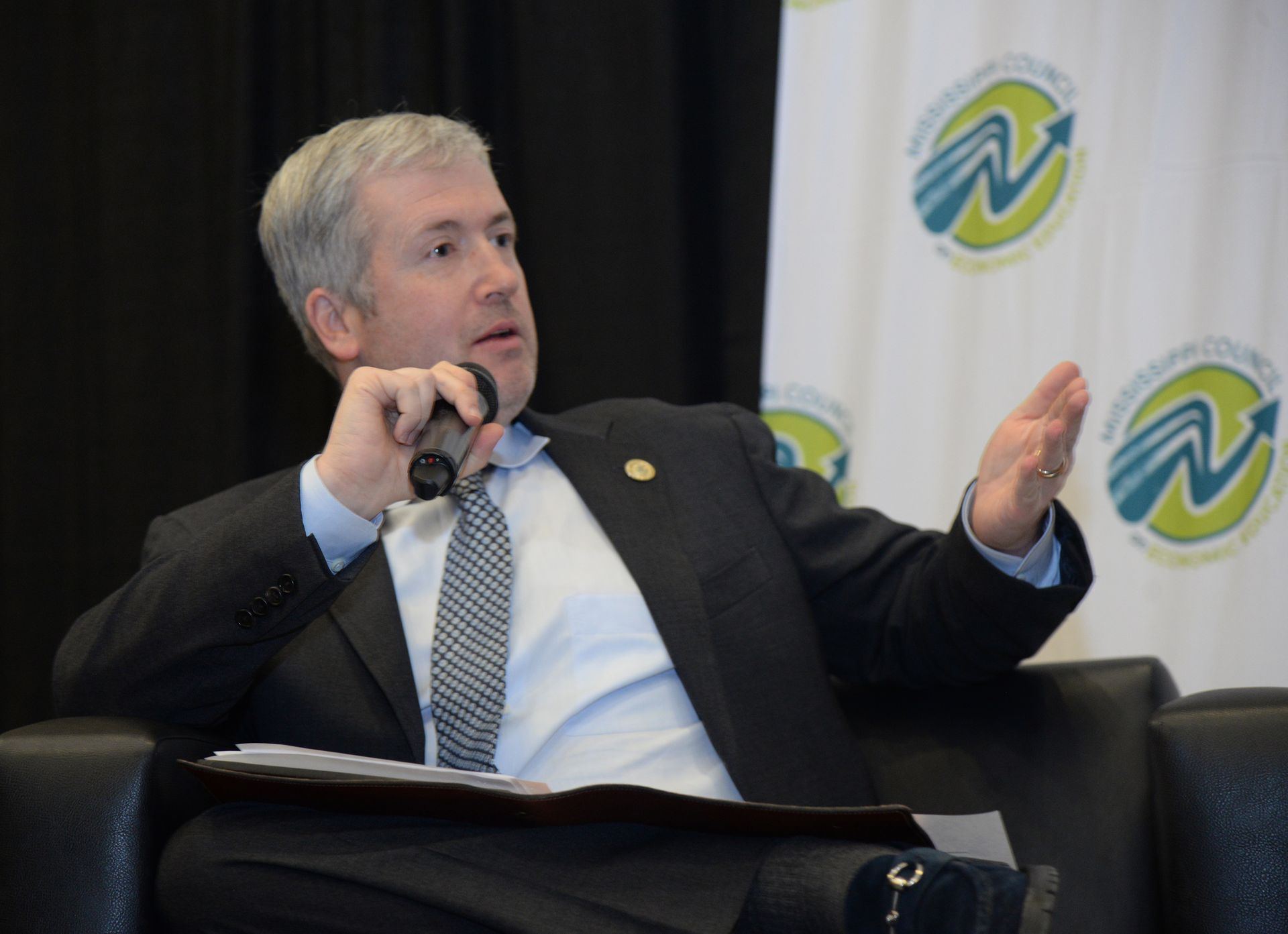 Man in a suit speaking into a microphone, gesturing with his right hand. He is seated on a black chair.