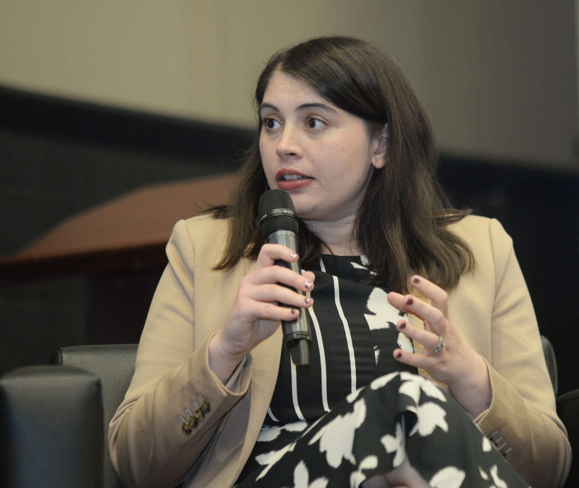 Woman speaking into a microphone, gesturing with hands; wearing blazer and black-and-white patterned dress.