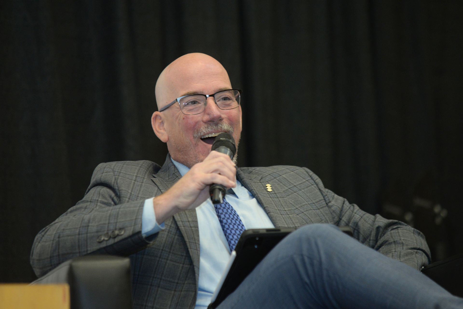 Man with glasses and bald head, holding a microphone and tablet, smiling while seated, dark backdrop.